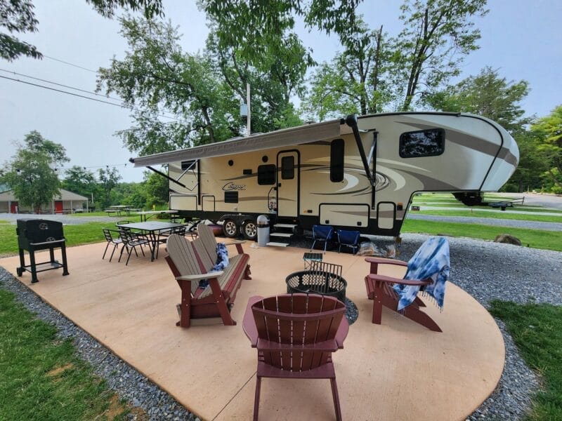 A fifth-wheel RV parked on a paved site with outdoor chairs, a fire pit, table, and a grill set up on a patio surrounded by grass and trees.