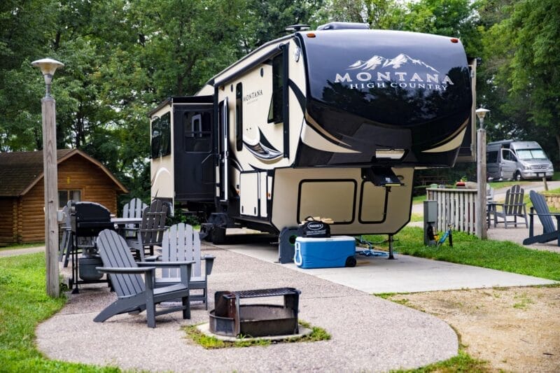 A large RV labeled "Montana High Country" is parked at a campsite with Adirondack chairs, a fire pit, and nearby trees.