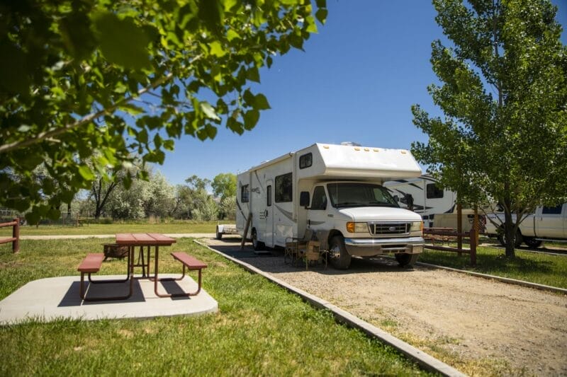 A white RV is parked in a campsite next to a picnic table under clear blue skies, with trees and grass surrounding the area.