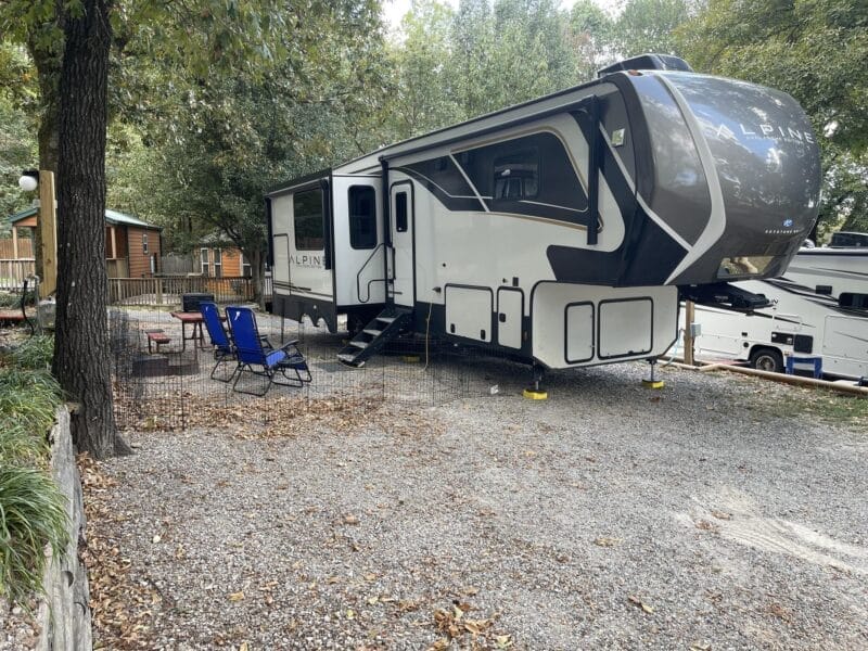 A large Alpine RV is parked on a gravel lot surrounded by trees, with outdoor chairs and a small fence set up beside it. A wooden cabin and another RV are visible in the background.
