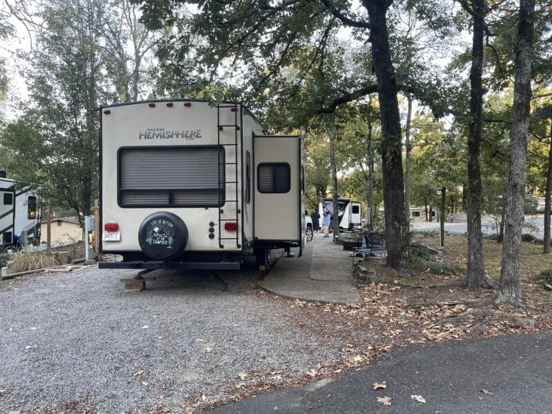 A parked travel trailer labeled "Salem Hemisphere" at a campsite surrounded by trees, with people and camping equipment visible in the background.