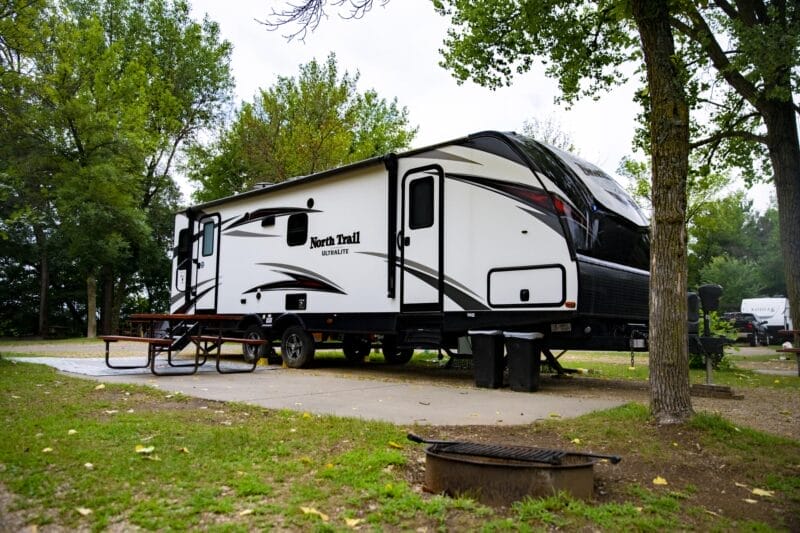 A white North Trail travel trailer is parked at a campsite with a picnic table, trees, and a fire ring nearby during daytime.
