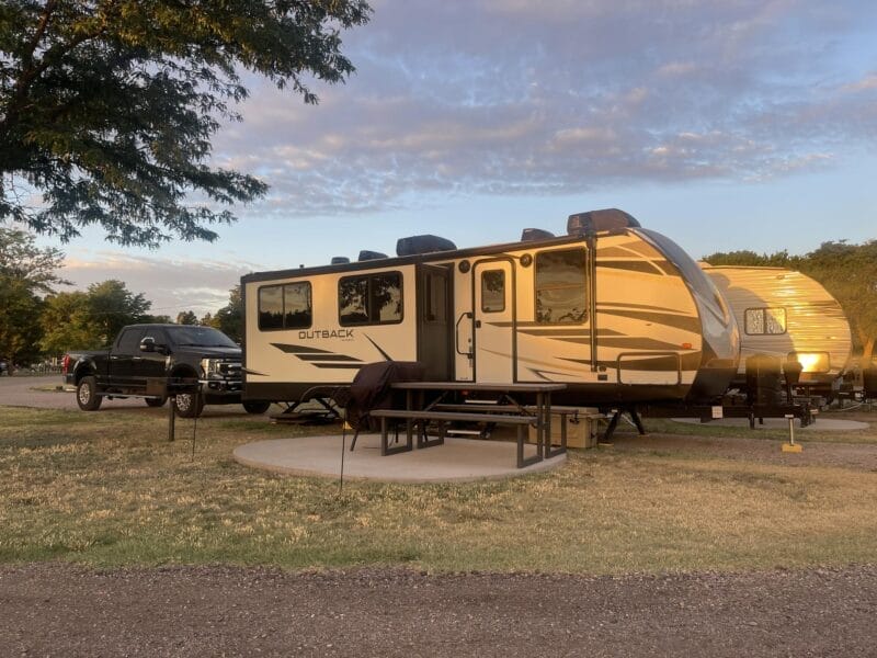 A beige and brown Outback travel trailer is parked at a campsite with a picnic table and a black pickup truck nearby under partly cloudy skies.