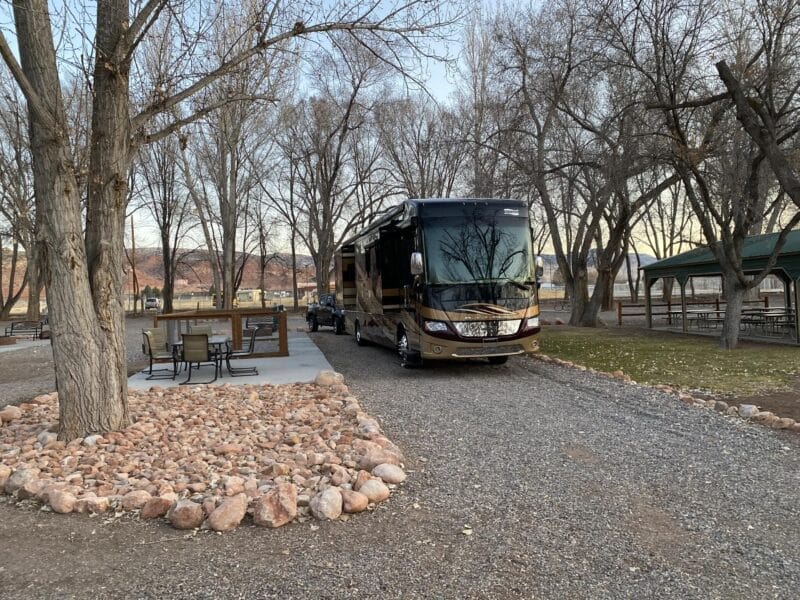A large motorhome is parked on a gravel campsite next to a picnic table and a fire pit, surrounded by leafless trees in a park setting.