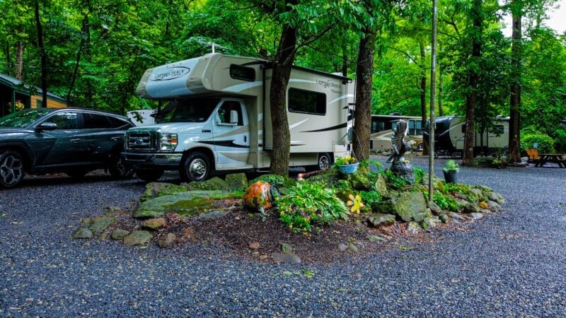 A white RV is parked on a gravel lot next to a garden area with rocks and decorative items, surrounded by trees and other vehicles.