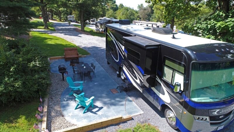 A large black and blue RV is parked at a campsite with a patio area featuring chairs, a table, and a bench surrounded by trees and greenery.
