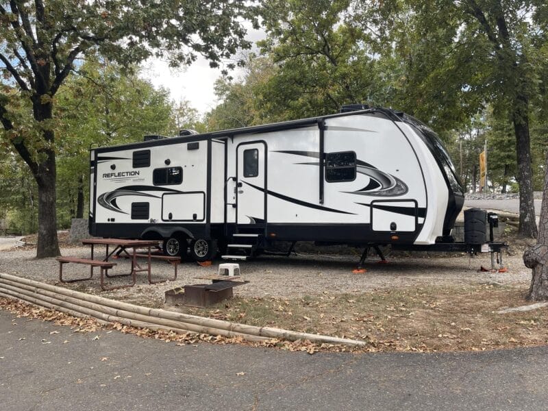 A white and black travel trailer is parked at a campsite with a picnic table, metal fire ring, gravel surface, and surrounding trees.