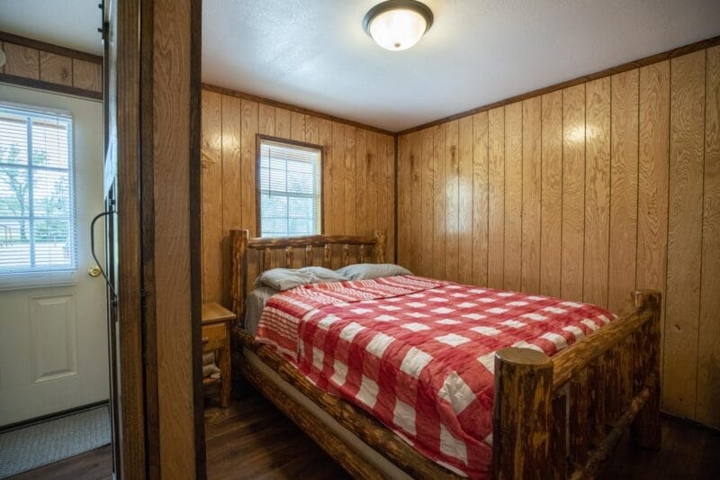 A small bedroom with wood-paneled walls features a rustic log bed with a red and white checkered quilt, a window, and a door leading outside.