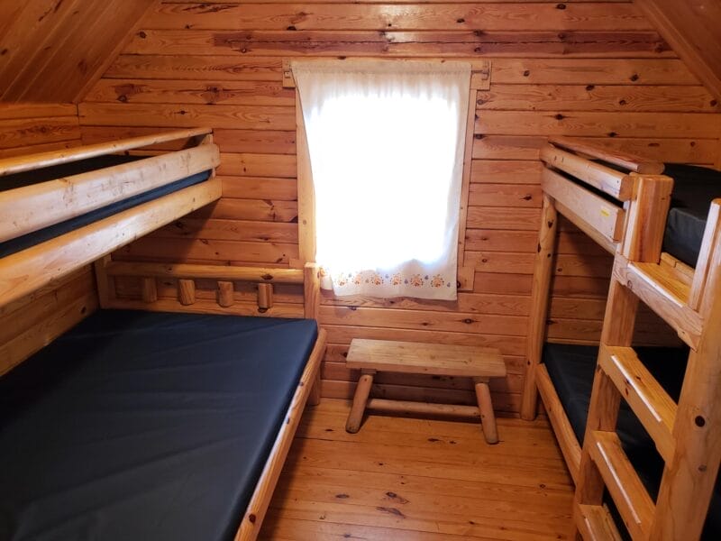 Wood-paneled room with two sets of wooden bunk beds, a small wooden bench, and a window with white curtains letting in natural light.
