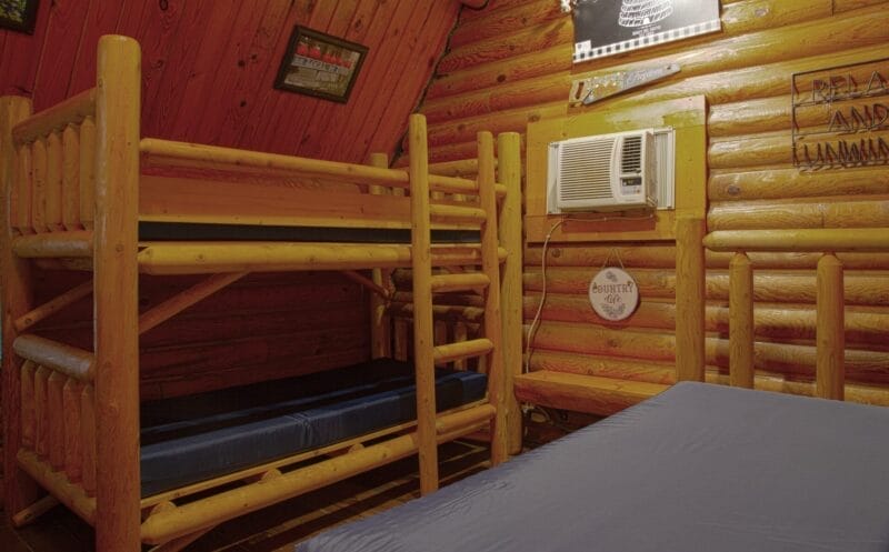A log cabin room with wooden bunk beds, a blue mattress, log walls, mounted air conditioner, and wall decorations.