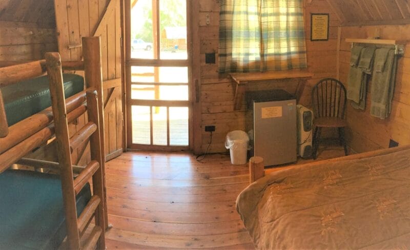 Small wooden cabin room with bunk beds, a double bed, a mini fridge, a window with plaid curtains, a chair, towels on the wall, and a door leading outside.