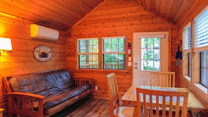 Small wooden cabin interior with wood-paneled walls, a futon, a dining table with chairs, windows, and a glass door leading outside.