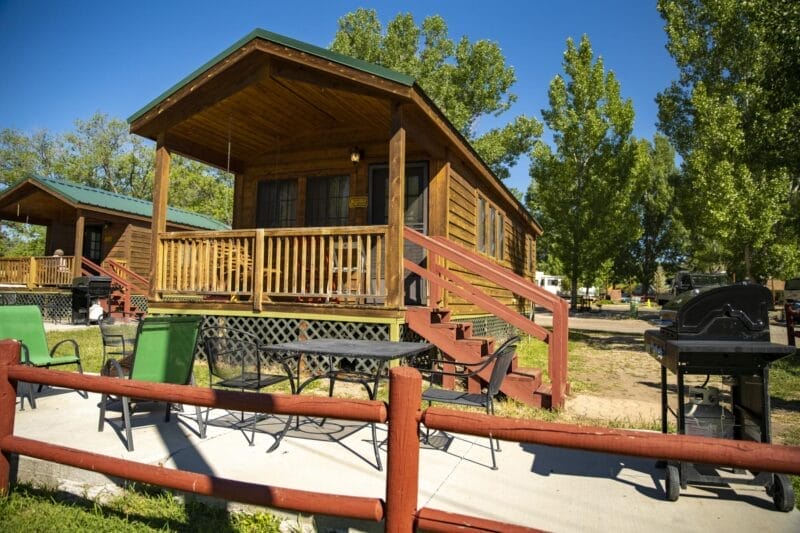 A small wooden cabin with a porch and green roof, outdoor seating, and grills, surrounded by grass, trees, and a red wooden fence on a sunny day.