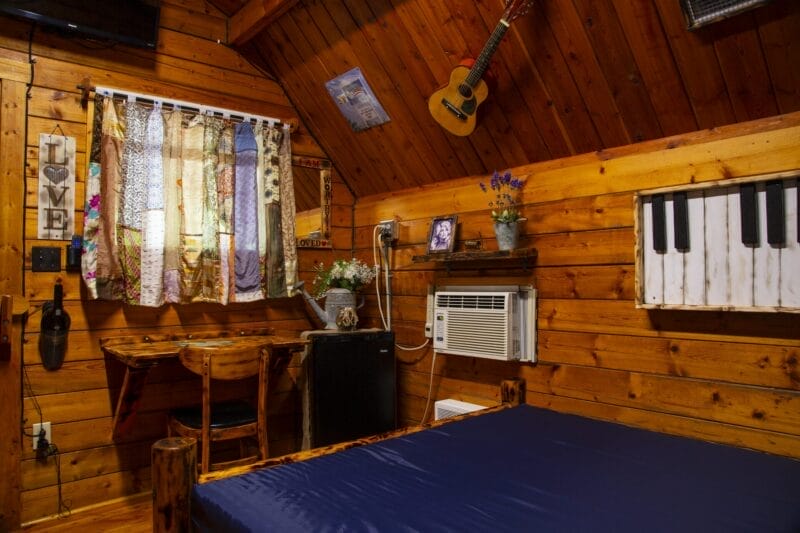 A cozy wooden cabin room features a bed, a small table with chairs, an air conditioner, wall-mounted guitar, and eclectic decorations including a piano key art piece and lace curtains.