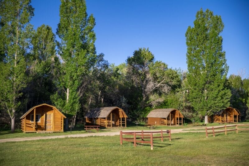 Four small wooden cabins with porches are spaced along a dirt path, surrounded by green grass, tall trees, and clear blue sky. Picnic tables and wooden fences are also visible.
