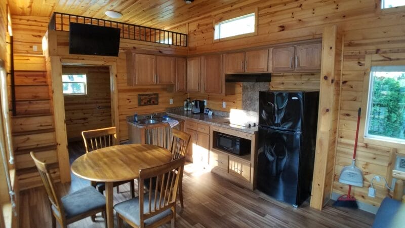 A small cabin kitchen with wood-paneled walls, a round dining table with four chairs, black appliances, and natural light from multiple windows.