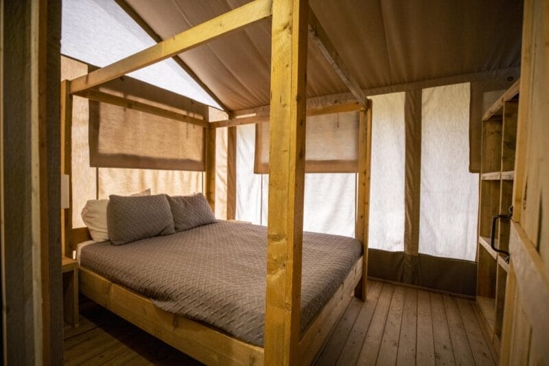 A wooden canopy bed with gray bedding is placed in a tent-like room with wooden floors and natural light coming through fabric walls.