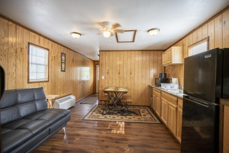 A small kitchen and living area features wood-paneled walls, a black sofa, dining table, black refrigerator, microwave, coffee maker, sink, and overhead lights.