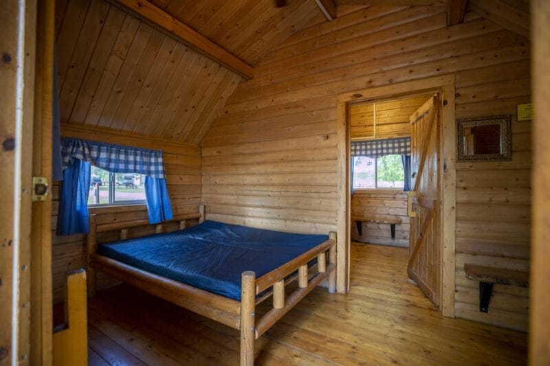A small wooden cabin room with a simple bed, blue curtains, wood-paneled walls, an open door, and a window showing greenery outside.