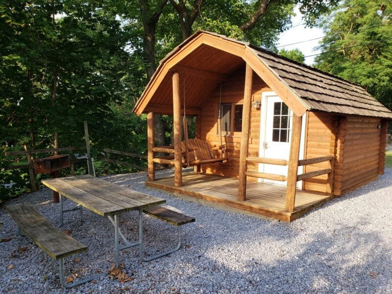 A small wooden cabin with a front porch swing, a white door, and a picnic table on gravel, surrounded by trees.