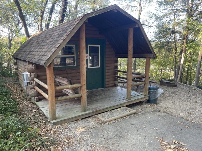 A small log cabin with a covered porch, wooden bench, and picnic table, surrounded by trees and fallen leaves.