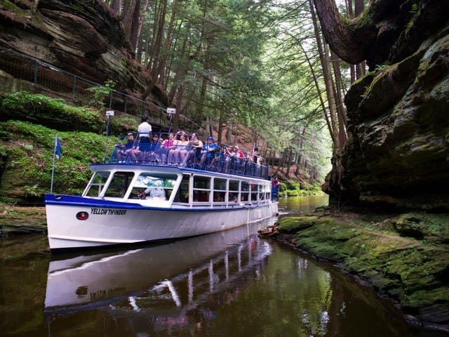 A white tour boat carrying passengers travels through a narrow, forested canyon with rocky cliffs and calm water.