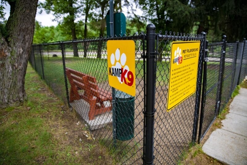 A fenced dog park area with a "Kamp K9" sign and pet playground rules posted on the gate, next to a sidewalk and trees.