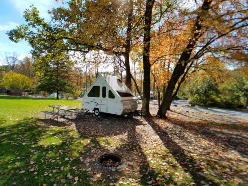 A small white pop-up camper is parked under trees with autumn leaves beside a picnic table in a grassy area near a river.