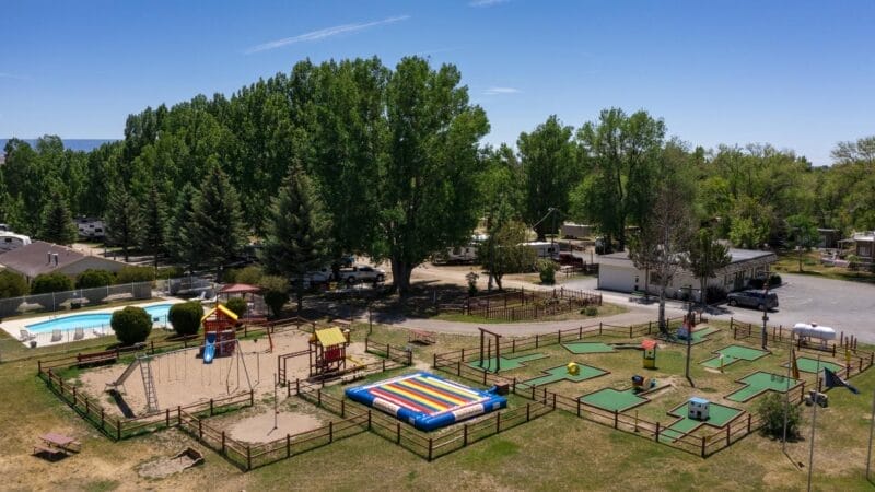Aerial view of a playground with swings, slides, a colorful inflatable, mini-golf course, and a swimming pool, surrounded by trees and nearby buildings.