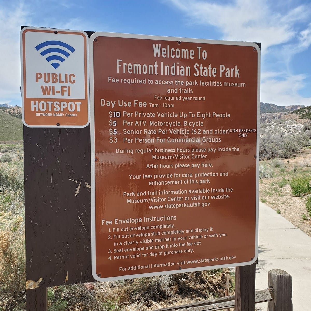 Brown sign at Fremont Indian State Park lists day use fees, visitor info, public Wi-Fi hotspot, and instructions for envelope submission. Desert landscape and pathway visible in background.