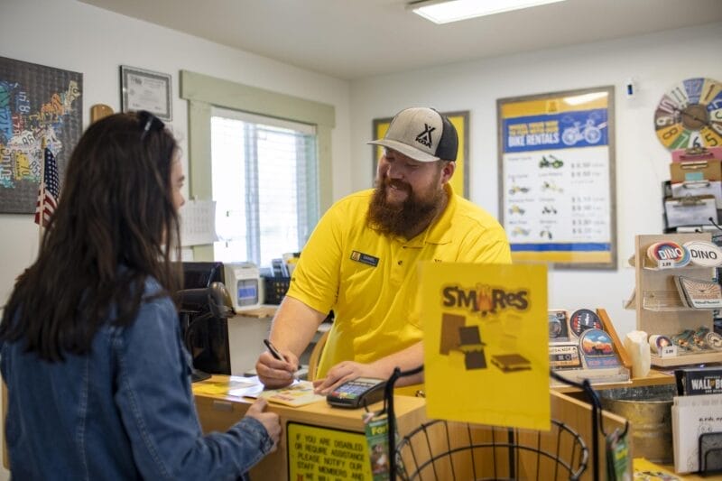 A man in a yellow shirt assists a woman at a front desk with informational signs, brochures, and a candy display visible on the counter.