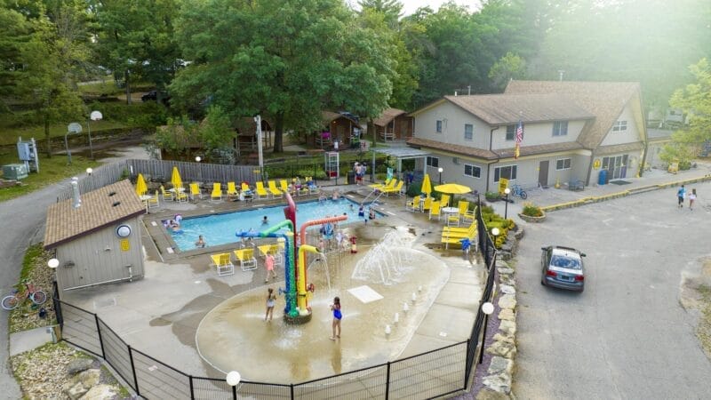 Aerial view of a community pool area with a splash pad, children playing, yellow lounge chairs, a building nearby, and trees surrounding the scene.