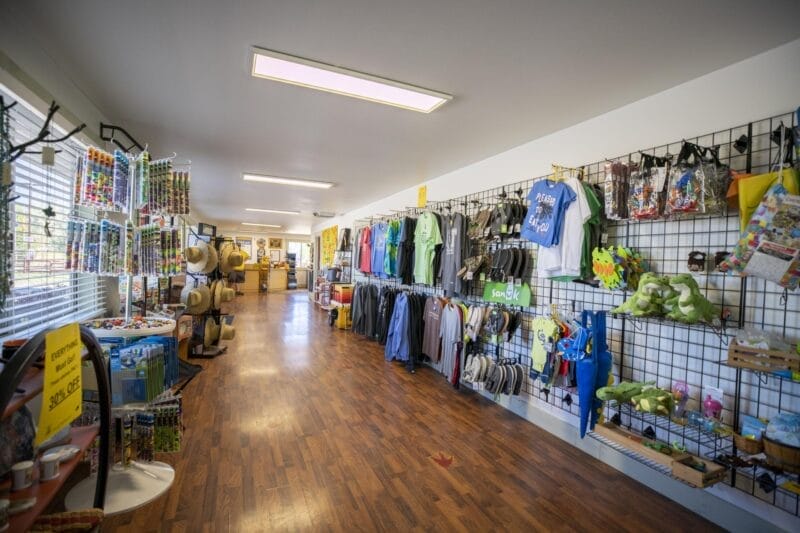 A gift shop with wooden floors, racks of hats, clothes, toys, and souvenirs displayed along the walls under bright ceiling lights.