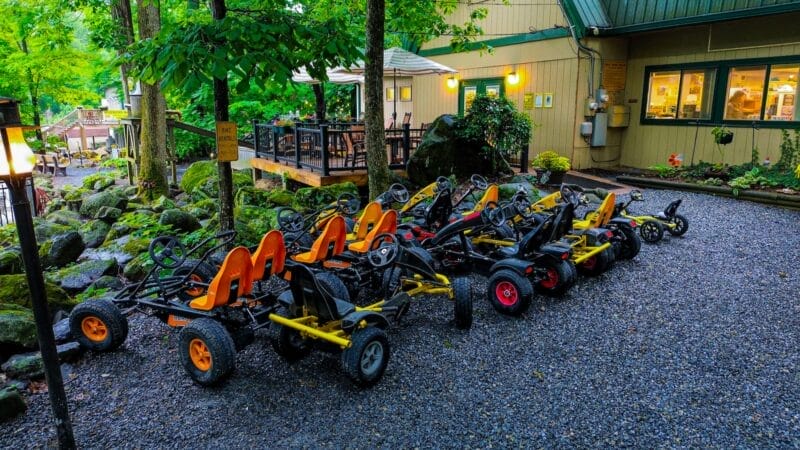 A row of pedal go-karts with orange and yellow seats is parked on a gravel area in front of a building surrounded by trees.