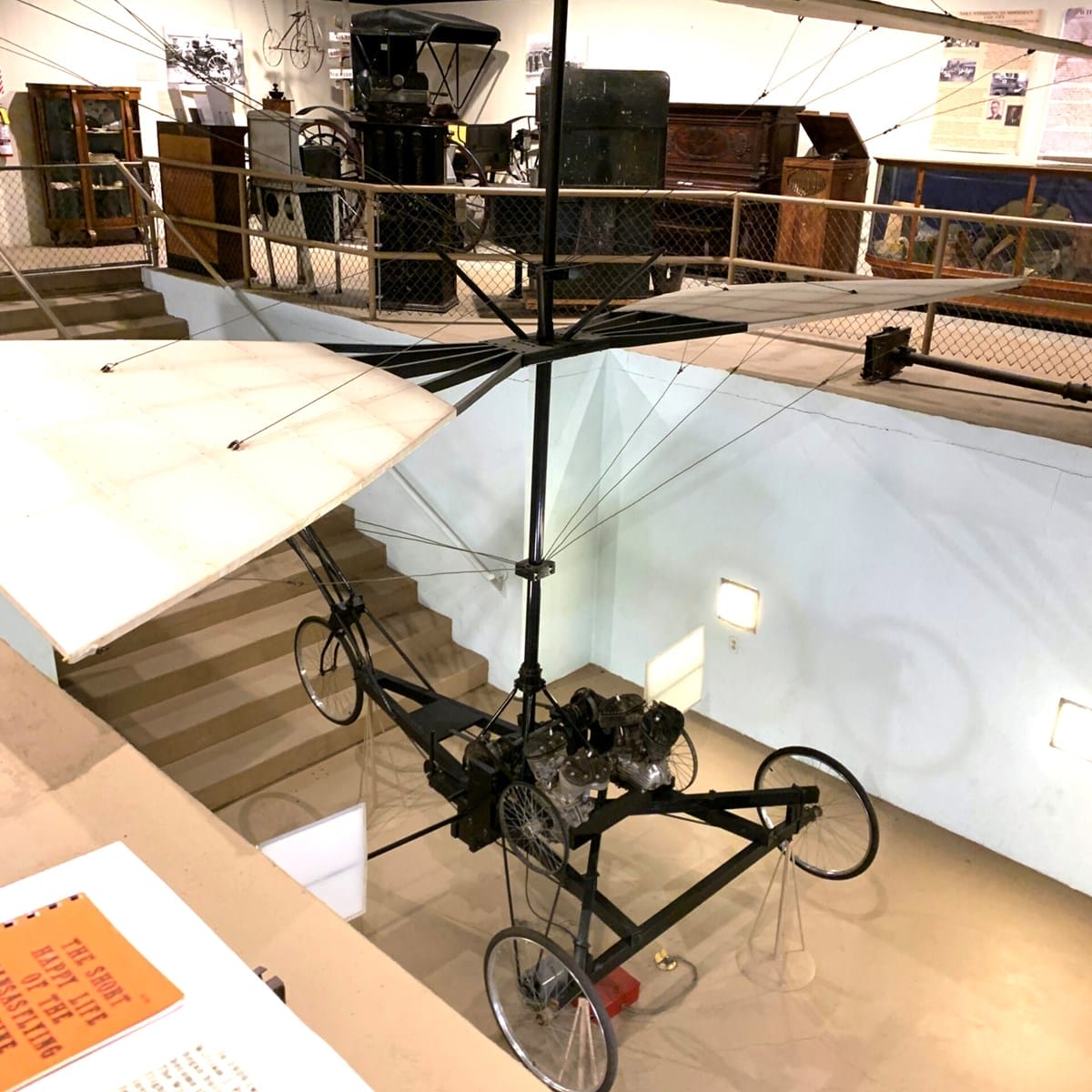 A vintage experimental aircraft with large wings and exposed framework is displayed indoors near a staircase at a museum.
