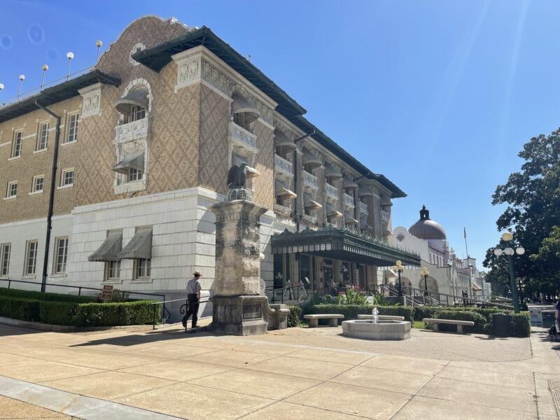 A historic multi-story building with balconies and columns, featuring a green canopy over the entrance and a small fountain in front, under a clear blue sky.