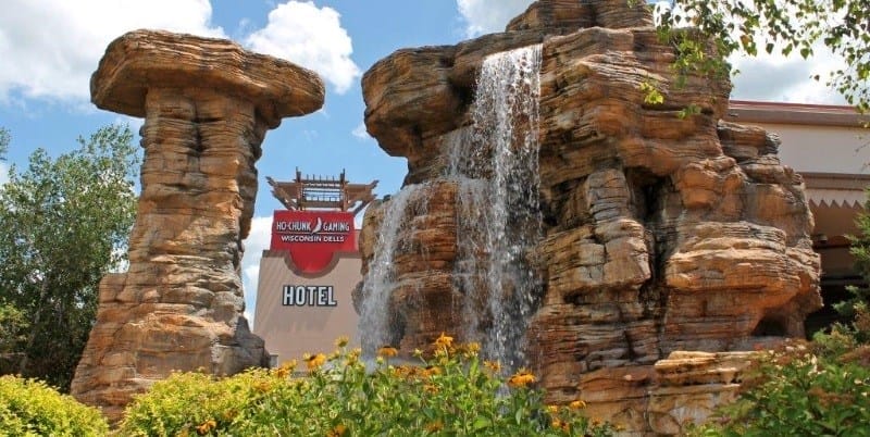 A man-made rock formation with a waterfall in front of a hotel sign that reads “Wisconsin Dells Hotel.” Flowers and greenery are in the foreground.