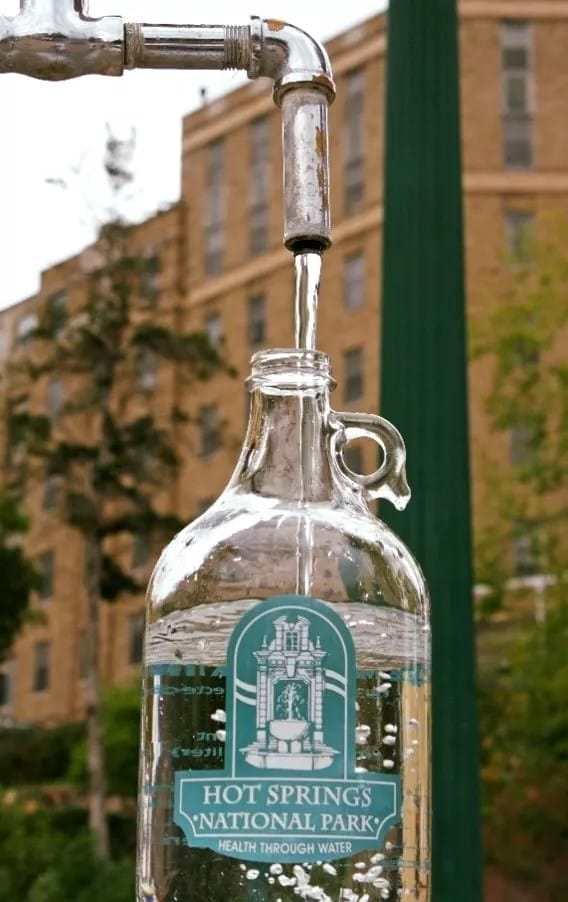 A glass jug labeled "Hot Springs National Park" is being filled with water from a metal spout, with buildings in the blurred background.