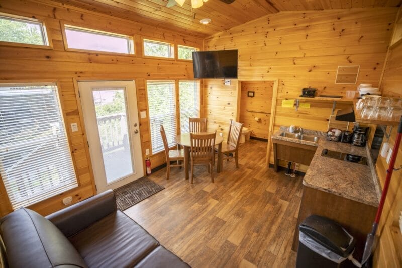 A small cabin interior with wooden walls, a sofa, a dining table with four chairs, a mounted TV, kitchen appliances, and large windows letting in natural light.