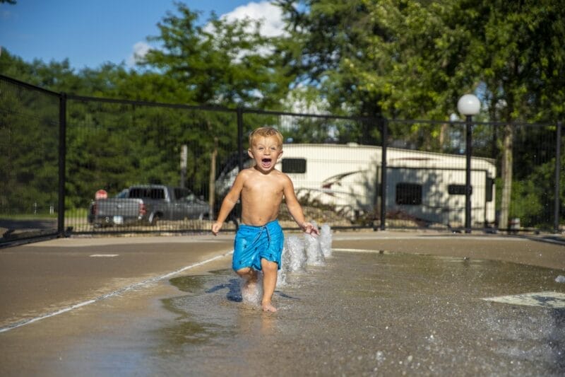 A young boy in blue swim trunks runs through a splash pad on a sunny day, with water jets spraying and trees and an RV in the background.