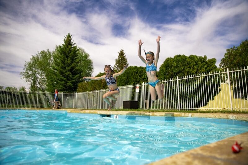 Two children in swimsuits are jumping into an outdoor swimming pool, with trees and a fence in the background under a partly cloudy sky.