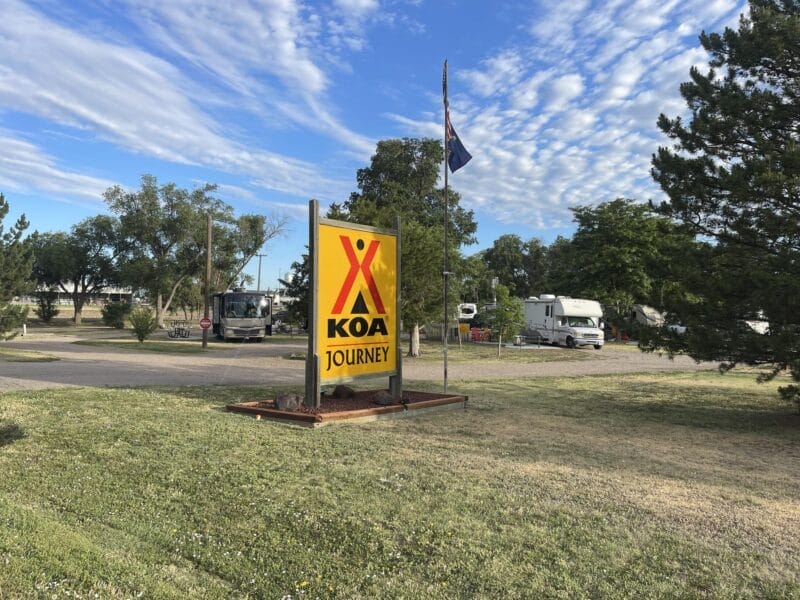 A KOA Journey campground sign stands near an American flag, trees, RVs, and a paved road under a partly cloudy sky.