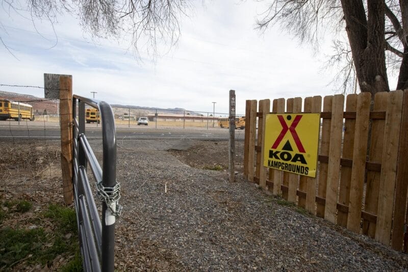 A KOA Kampgrounds sign is attached to a wooden fence beside a gate, with a gravel path leading to a road and school buses visible in the background.