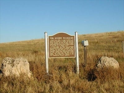 A historical marker titled "The Kidder Massacre" stands in a grassy field between two rocks and next to a small gray mailbox under a clear blue sky.