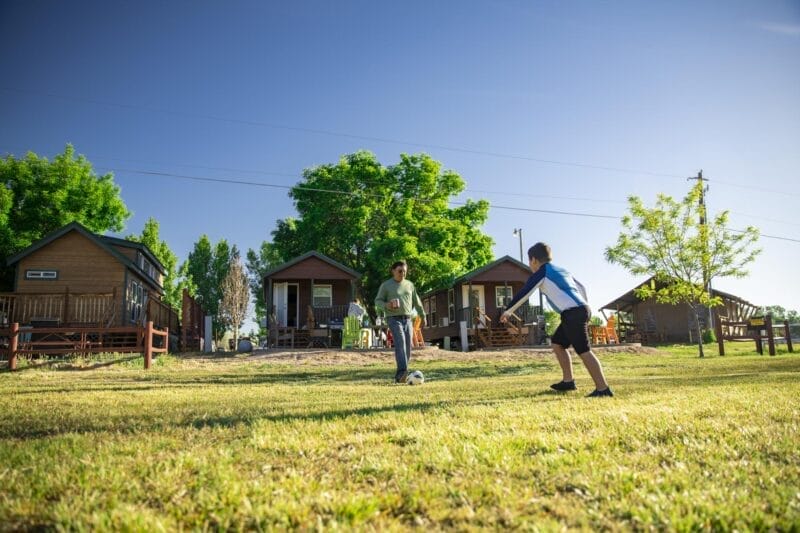 Two children play soccer on a grassy field in front of several small wooden cabins under a clear blue sky.