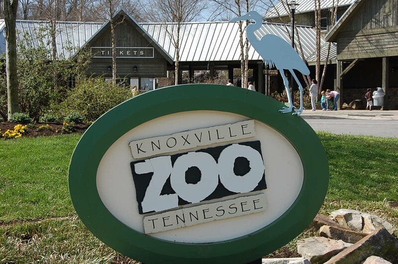 Knoxville Zoo entrance sign with a bird sculpture on top, ticket booth and visitors in the background.