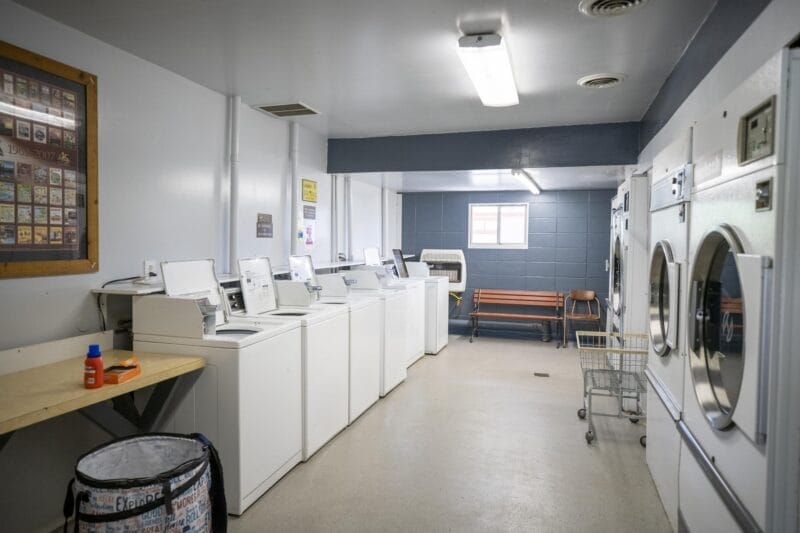 A clean, well-lit laundromat room with several washing machines and dryers, a folding table, laundry supplies, a bench, a laundry cart, and a small window.