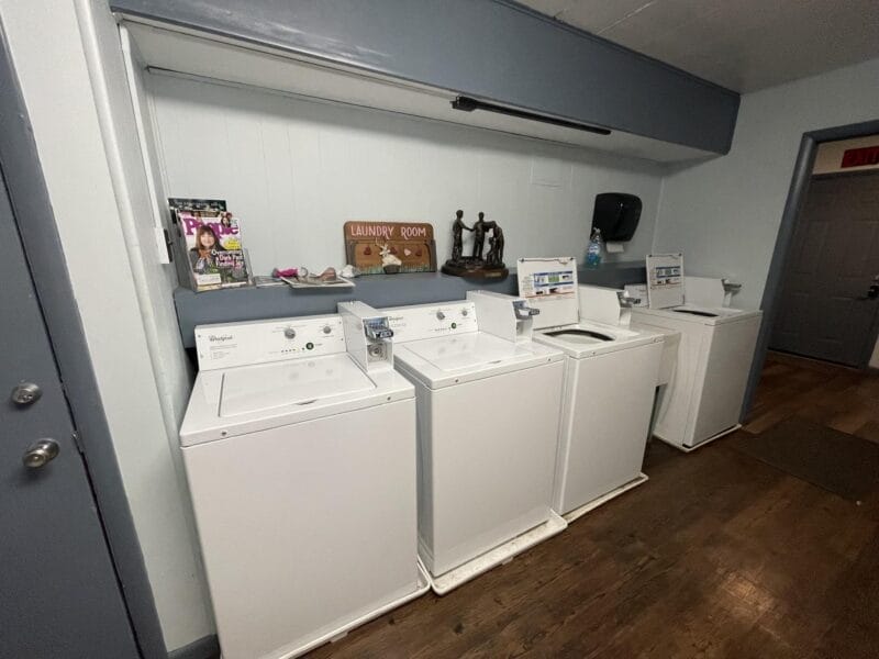 Four white washing machines and dryers are lined up against a wall in a laundry room with a wooden floor and various items on the shelf above.