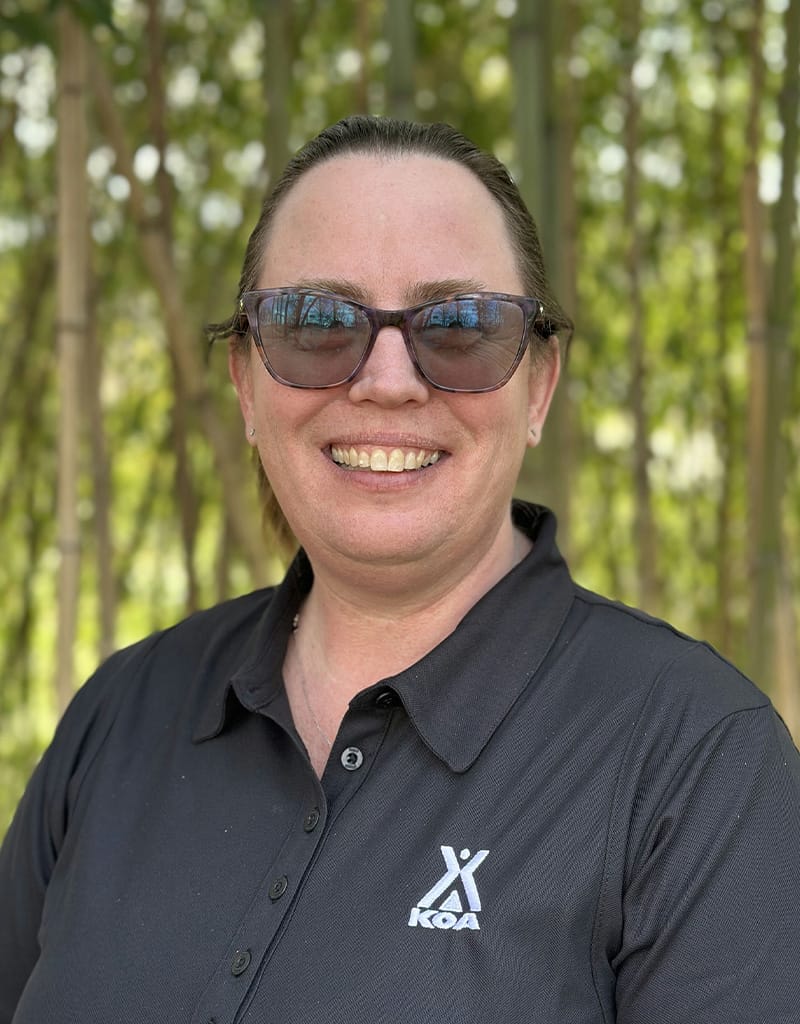 Lee-Ann-Oliver is wearing sunglasses and a black KOA polo shirt, smiling outdoors.