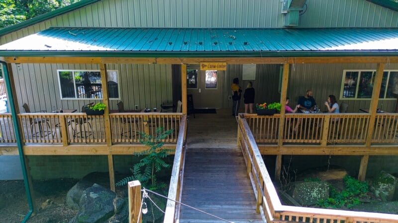 A wooden ramp leads to the entrance of a building with a green metal roof. Several people are sitting or standing on the porch. The sign above the door reads “Dining Lodge.”.